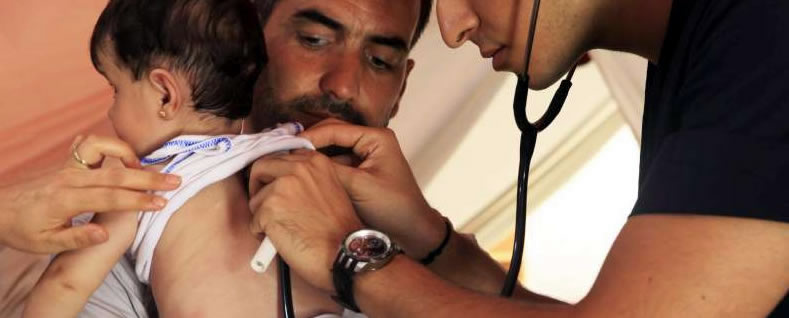 A father holds his young child as medical staff listen to the childs chest